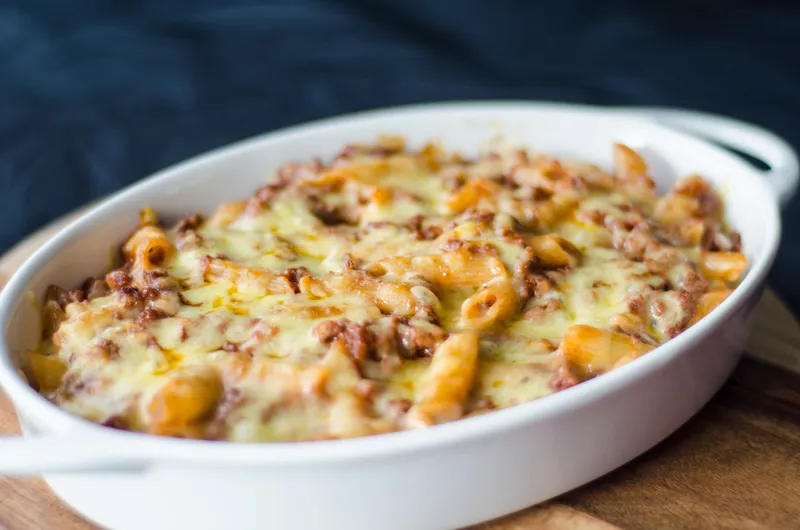 Nudelauflauf mit Hackfleisch und überbackenem Käse in einer ovalen weißen Auflaufform auf Holzbrett