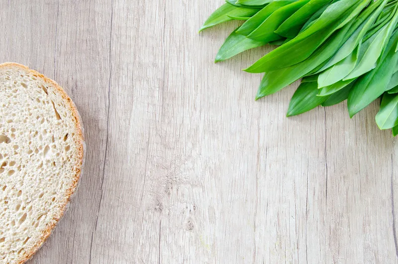Ein Bund frischer Bärlauchblätter und eine Scheibe Brot liegen auf einer hellen Holzoberfläche.