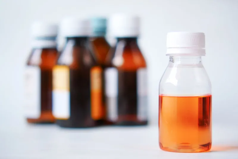 Mehrere braune Medikamentenfläschchen mit weißen Verschlüssen, davor eine durchsichtige Flasche mit orangefarbenem Saft