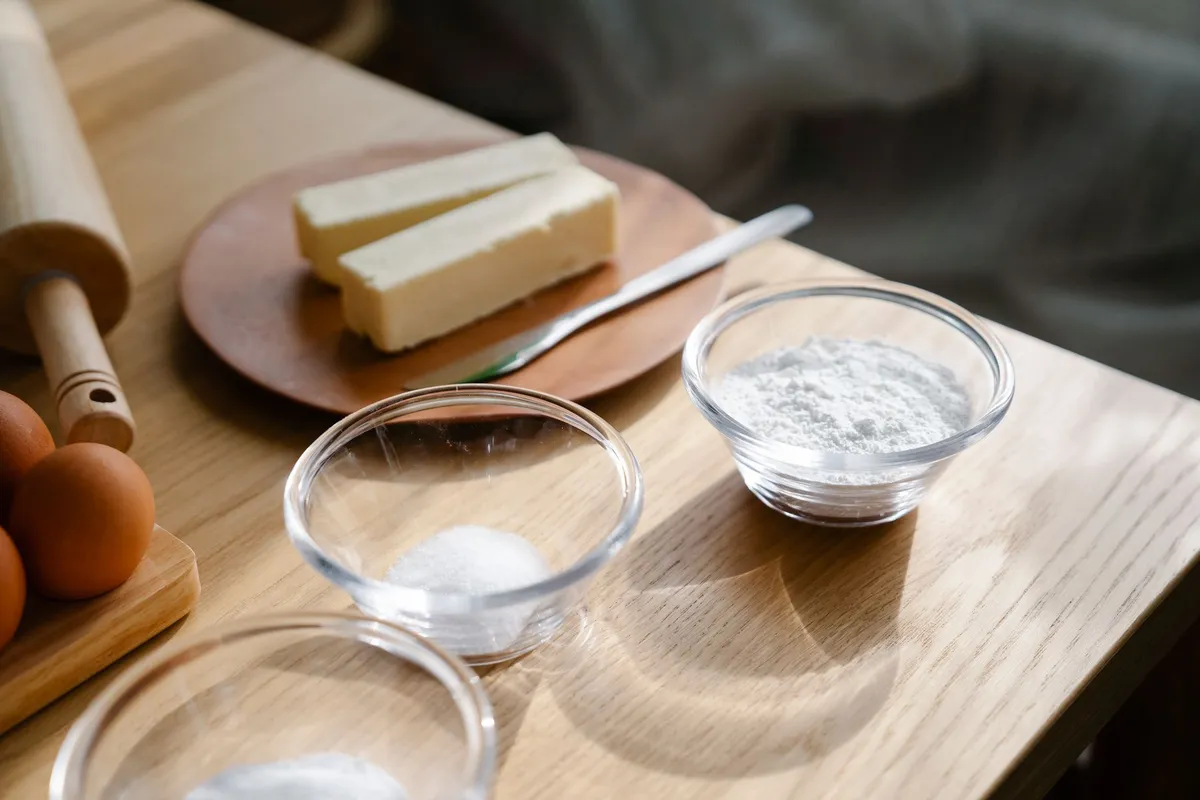 Zutaten für Butterplätzchen auf einem Holztisch: Butterstücke, Mehl/Zucker, ein Ei und eine Teigrolle.