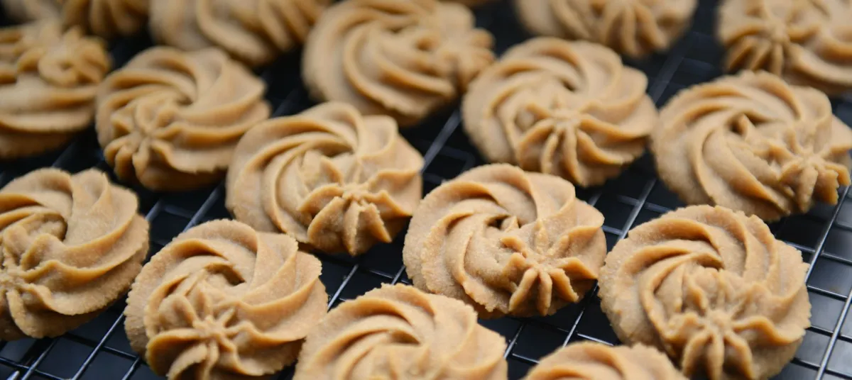 Zahlreiche goldbraune, spiralförmige Butterplätzchen kühlen auf einem schwarzen Gitterrost ab.