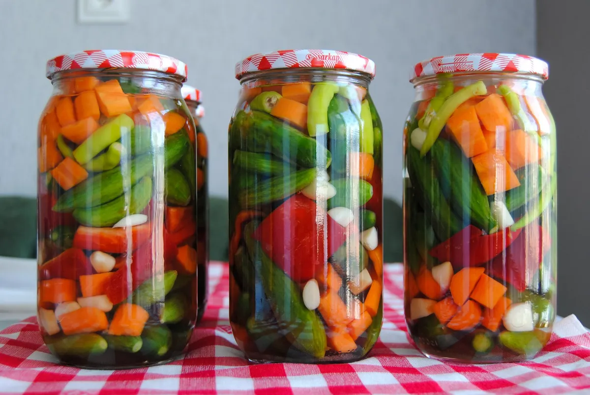 Drei Gläser mit eingelegtem Gemüse aus Karottenwürfeln, Gurken, Paprika und Knoblauch in Lake, auf kariertem Tuch