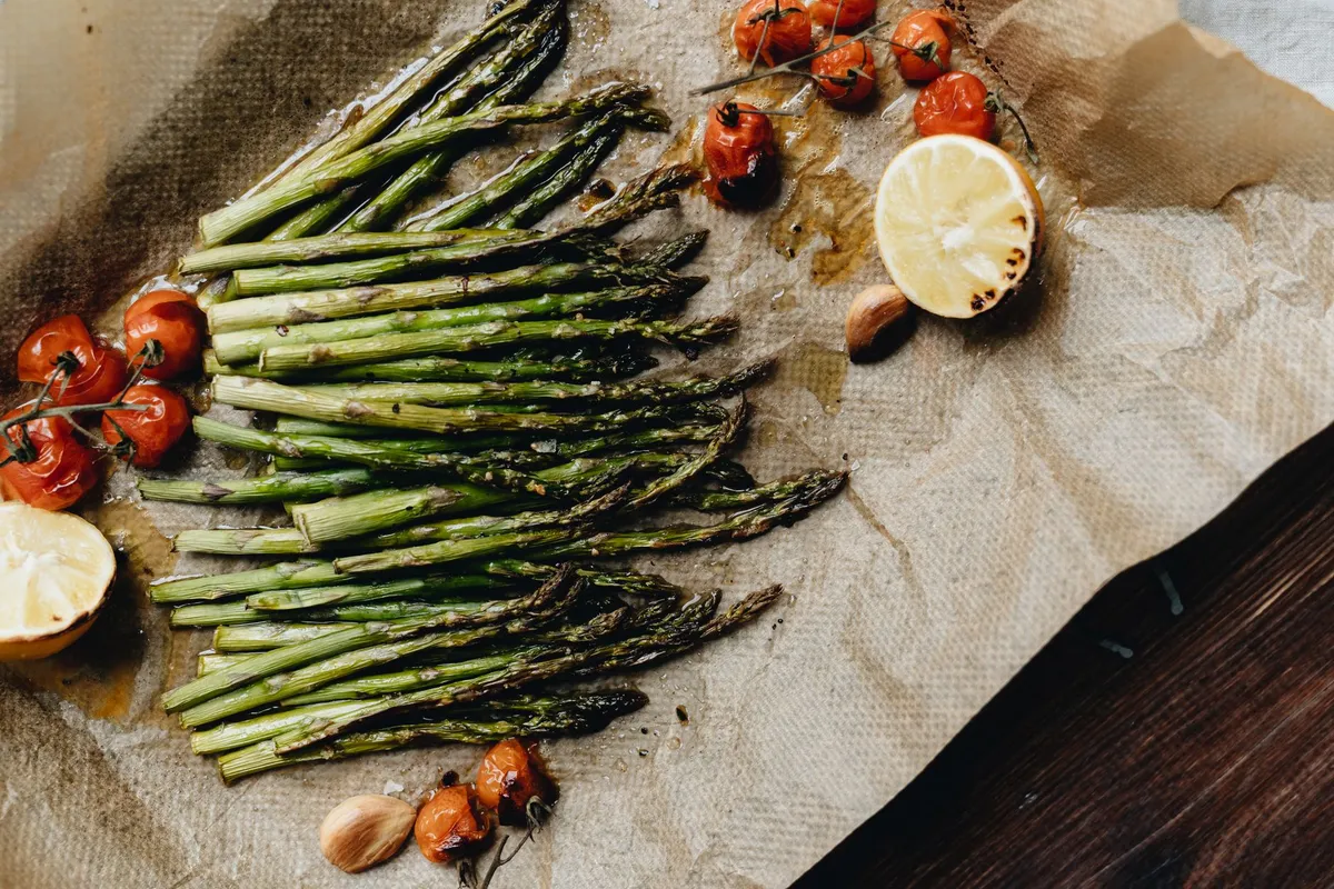 Gebackener grüner Spargel auf Backpapier, daneben Kirschtomaten, Zitronenhälften und Knoblauchzehen.