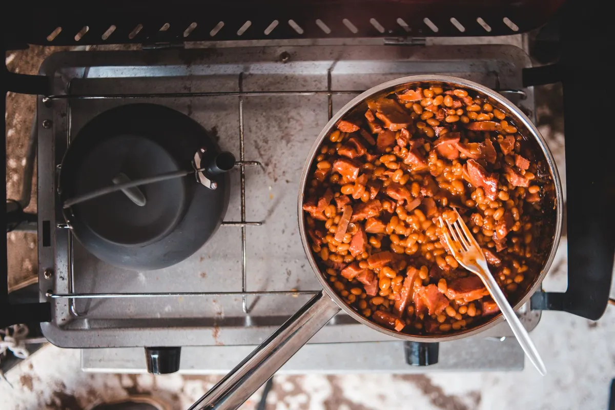 Pfanne mit Bohnen in Tomatensauce und Speckstreifen auf einem Campingkocher, darin steckt eine Gabel