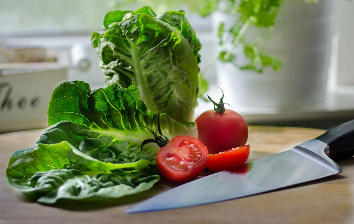 Frische grüne Salatblätter, eine ganze und eine geschnittene Tomate sowie ein Messer liegen auf einem Holzbrett.