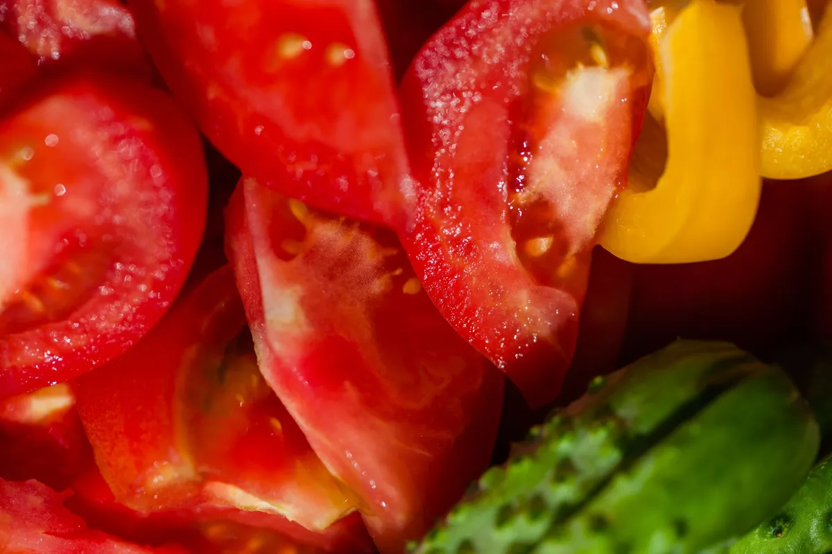 Grob geschnittene Tomatenstücke und gelbe Paprikastücke in Nahaufnahme, daneben eine grüne Gurke.