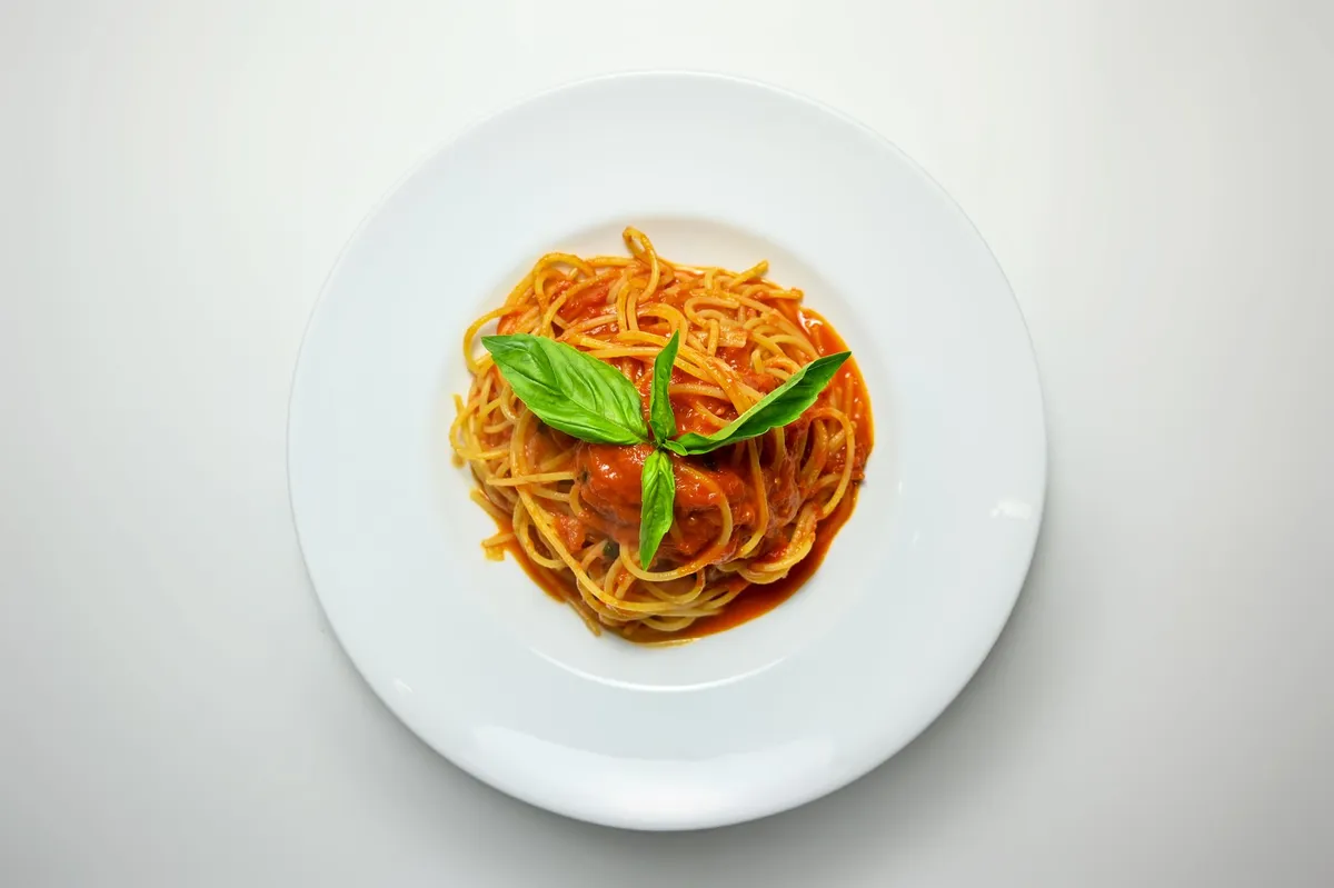 Spaghetti mit Tomatensoße und drei Basilikumblättern auf einem weißen Teller, von oben fotografiert.