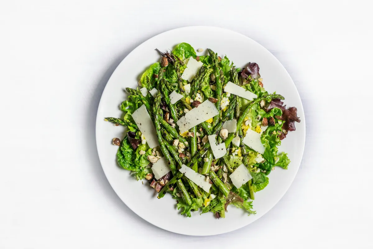 Spargelsalat mit grünem Spargel, Salatblättern und Hobelkäse auf einem weißen Teller