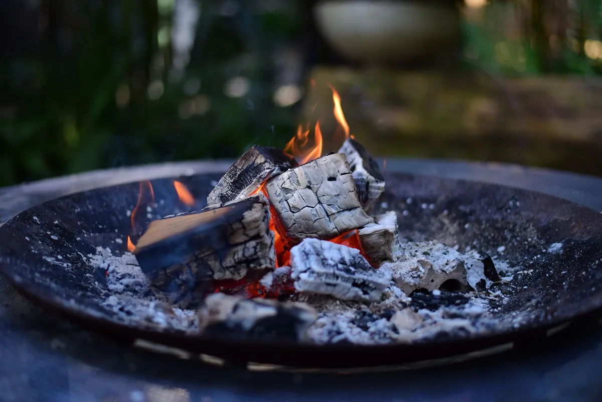 Glühende Holzscheite in einer sicheren Feuerschale im Garten bei Dämmerung.