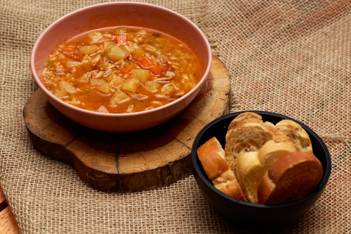 Suppe mit Kartoffelwürfeln, Karotten und Getreide in rosa Schale, daneben eine Schale mit Brotscheiben