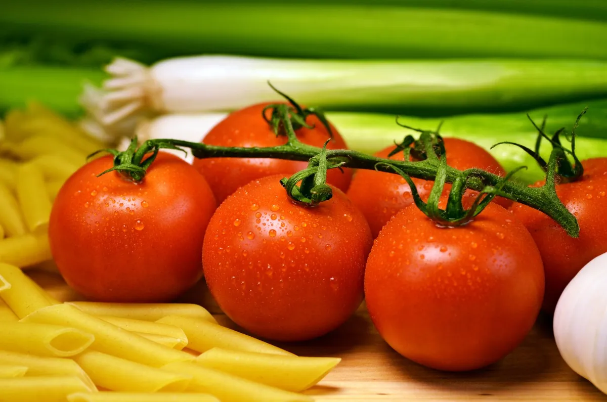 Frische Strauchtomaten mit Wassertropfen auf Holz, daneben Frühlingszwiebeln, Knoblauch und gelbe Nudeln.