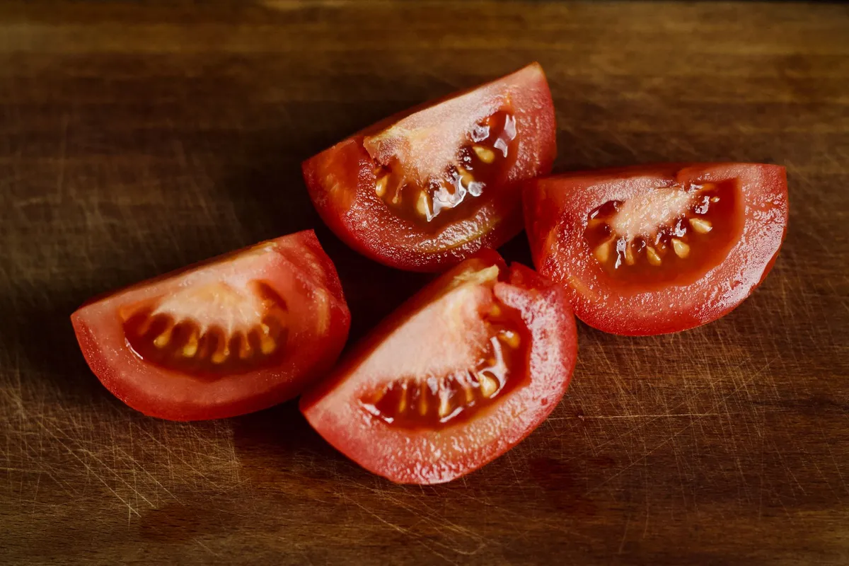 Vier Tomatenviertel mit sichtbaren Kernen liegen auf einem dunklen, verkratzten Holzbrett.