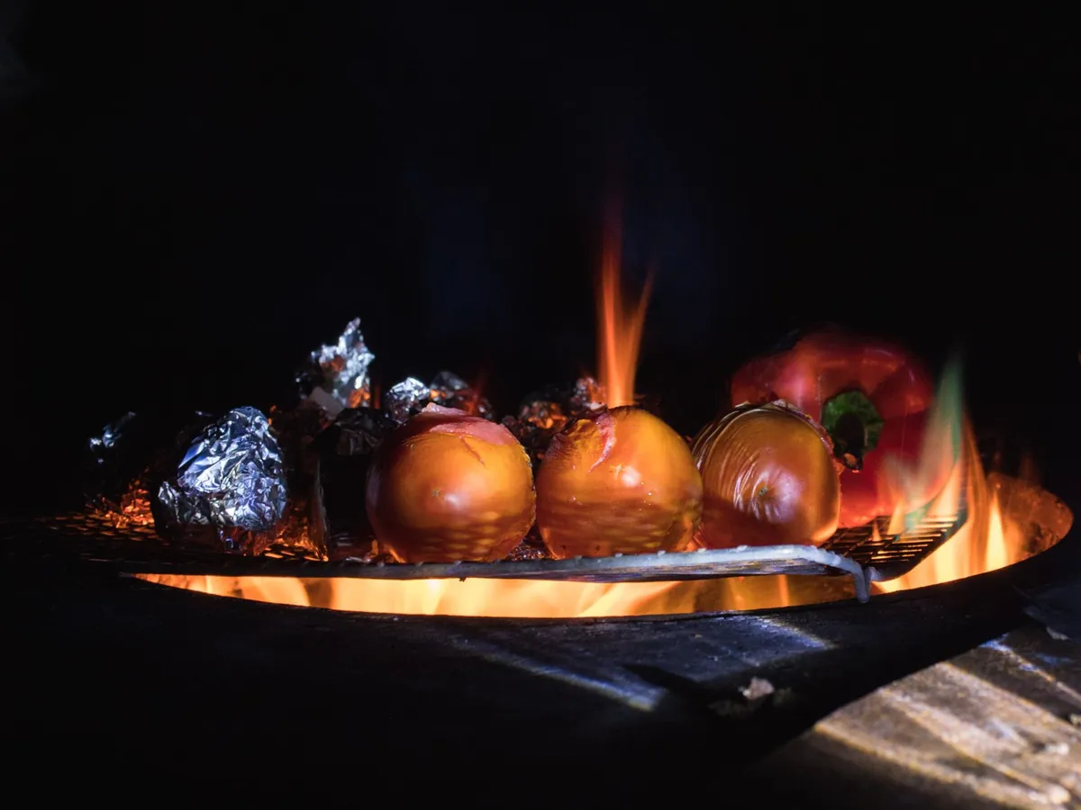 Ganze Tomaten und eine rote Paprika liegen neben Alufolienpaketen auf einem Grill über offener Flamme.