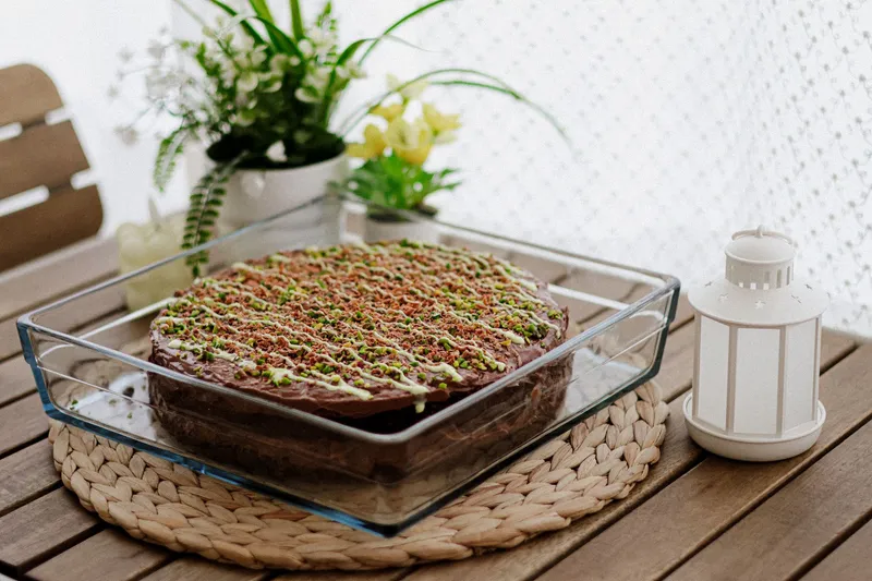 Schokoladenkuchen mit Pistazien und hellem Guss in einer eckigen Glasform auf einem Holztisch