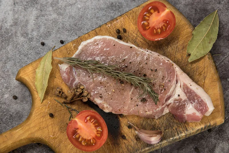 Rohes Fleisch, Rosmarinzweige, Tomatenhälften und Gewürze liegen auf einem rustikalen Holzschneidebrett.