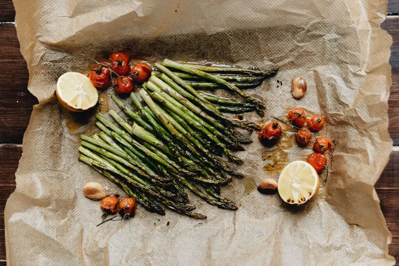 Gebratener grüner Spargel auf Backpapier mit Kirschtomaten, Zitronenhälften und Knoblauchzehen