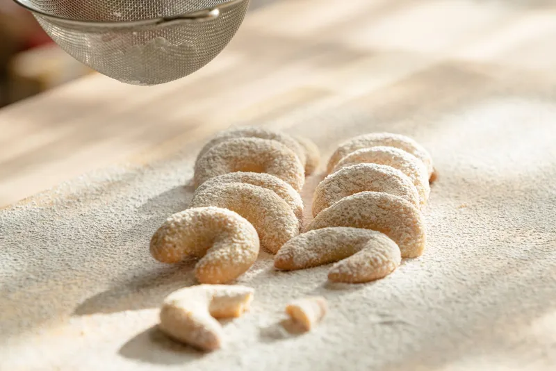 Mehrere Vanillekipferl liegen auf einer Holzoberfläche und werden mit Puderzucker bestreut.